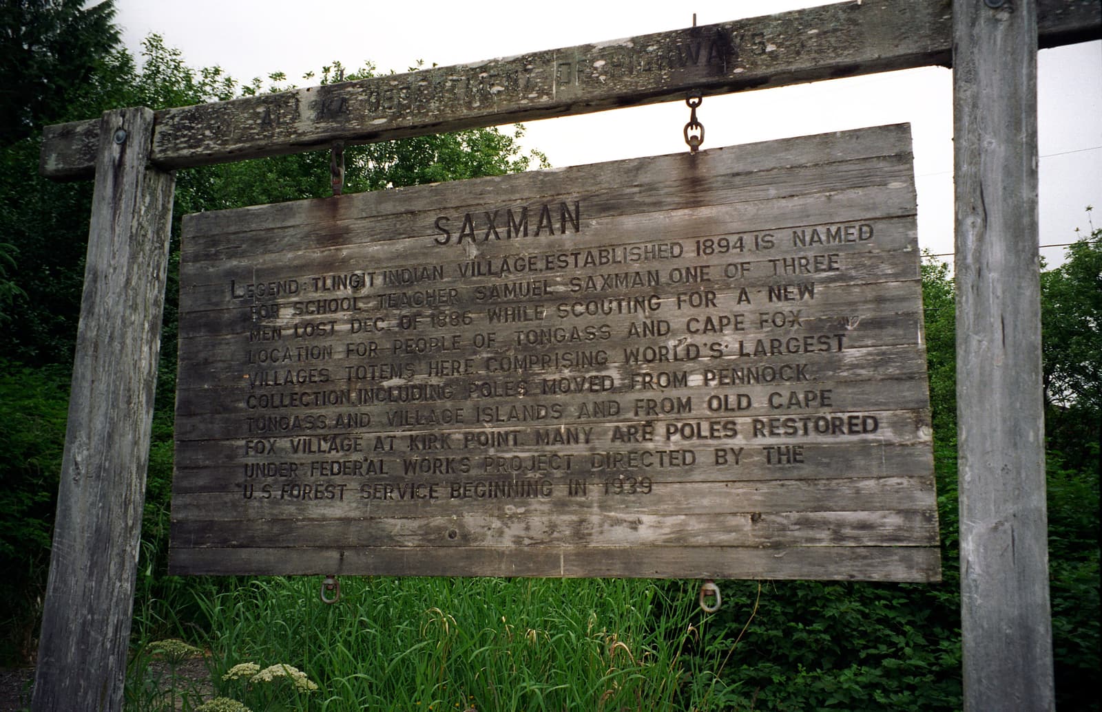 A weathered wooden sign recounting Saxman’s history at the totem park